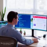 Web developer coding a crypto security platform on dual monitors in a modern office – The Investors Centre branding visible