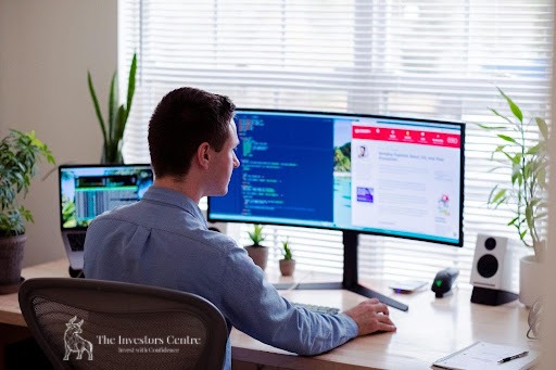 Web developer coding a crypto security platform on dual monitors in a modern office – The Investors Centre branding visible